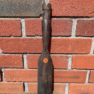 May include: A dark brown, weathered metal chisel with a long, slender handle and a wide, flat blade. The tool features a cylindrical top section and a distinct, tapered neck leading to the broad cutting edge. An orange circular sticker with the number 140 is affixed to the center of the blade.