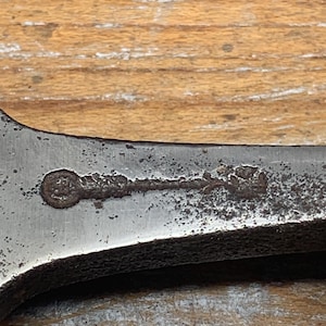 May include: Close-up of a vintage, well-worn wrench. The metal tool shows signs of age with rust and wear. A small, circular marking is visible on the surface. The wrench is set against a wooden background.