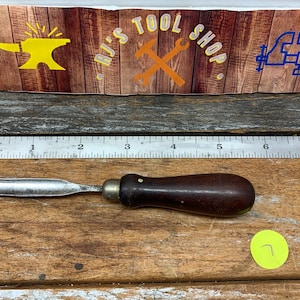 May include: A small, wooden-handled, metal carving tool with a rounded blade. The tool is about 6 inches long and has a dark brown handle. The tool is lying on a wooden surface.