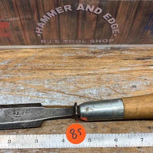 May include: A vintage chisel with a wooden handle and a metal blade. The blade is dark and shows signs of wear. The handle is light brown. The tool is placed on a wooden surface with a ruler.