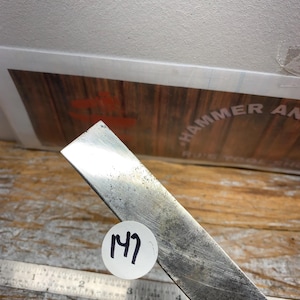 May include: A metal chisel with a worn, textured surface. A white sticker with the number "147" is affixed to the chisel. A metal ruler is visible in the background. The chisel is likely used for woodworking or metalworking.