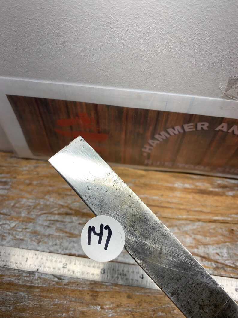 May include: A metal chisel with a worn, textured surface. A white sticker with the number "147" is affixed to the chisel. A metal ruler is visible in the background. The chisel is likely used for woodworking or metalworking.