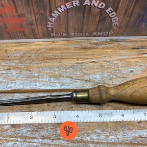 May include: A wood-handled chisel with a metal blade, brass ferrule, and a ruler in the foreground. The chisel is on a wooden surface. The background features the text "HAMMER AND EDGE" and "RJ'S TOOL SHOP".
