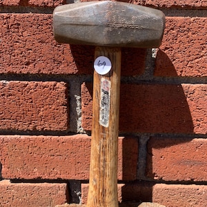 May include: A well-worn, metal-headed hammer with a wooden handle. The hammerhead is a dark, metallic gray, and the handle shows signs of use. A small, white sticker with the number "69" is affixed to the handle. The background is a red brick wall.