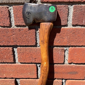 May include: A metal hatchet with a dark, weathered steel head featuring the handwritten number 1.9 and a green circular sticker with the number 91. The hatchet has a smooth, light brown wooden handle with a visible grain and a single metal pin.
