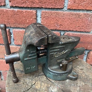 May include: A vintage green metal bench vise with a rotating base and a steel handle. The side of the vise features the embossed text "Chas" and "No 44". The tool has a weathered, industrial appearance with visible signs of use and oxidation.