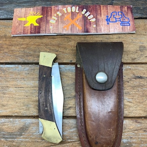 May include: A folding knife with a brown wood handle and a brass bolster. The knife has a stainless steel blade and comes with a brown leather sheath.