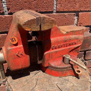 May include: A red Craftsman bench vise with a metallic handle and a rusted top surface. The tool features a prominent orange circular sticker with the number 81 printed in black. The body of the vise is cast metal with visible wear and oxidation.