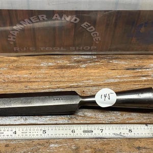 May include: A metal chisel with a wooden handle, resting on a wooden surface. The chisel has a dark, rectangular blade and a light-colored, cylindrical handle. A metal ruler is below the chisel. The text "HAMMER AND EDGE" is visible.