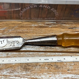 May include: A Craftsman wood chisel with a clear handle and a metal blade. A metal ruler is placed below the chisel. The chisel has a sticker with the number 125 on it. The background includes a sign that says "Hammer and Edge" and "NJ's Tool Shop."