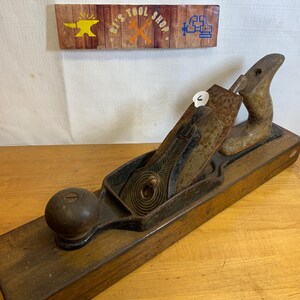 May include: A vintage wooden hand plane with a dark wooden body and a metal blade. The plane has a wooden handle and knob. Above the plane is a wooden sign that reads "Ri's Tool Shop."