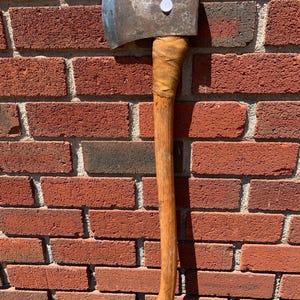 May include: A vintage axe with a wooden handle and a metal blade. The handle is wrapped with a tan material near the head. The axe is set against a red brick wall.