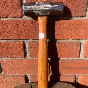 May include: A metal mallet with a wooden handle. The mallet head is silver and rectangular, with a slightly curved striking surface. The handle is made of light brown wood. A small white sticker with the number 50 is on the handle.