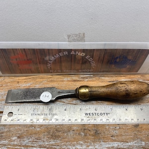 May include: A vintage chisel with a wooden handle and a metal blade. The chisel is placed on a wooden surface next to a stainless steel ruler. A sticker with the number 134 is on the chisel.