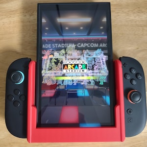 以下が含まれることがあります： 赤色のフレームと黒いコントローラーが付いた黒色の携帯ゲーム機。画面にはCapcom Arcade Stadiumのゲームインターフェースが表示されています。ゲームのロゴと「Start Game」の文字が表示されています。