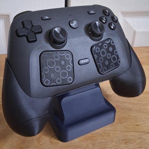 Può includere: Un controller per videogiochi nero opaco con un pulsante centrale con logo Steam, due levette analogiche, un pad direzionale e quattro pulsanti azione etichettati A, B, X e Y. Due trackpad quadrati con motivo circolare sotto le levette.