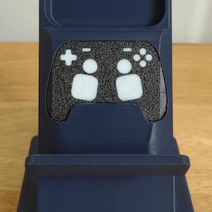 May include: A navy blue phone stand with a controller design. The stand has a textured black panel with white controller symbols. The stand is designed to hold a phone and has a base for stability.