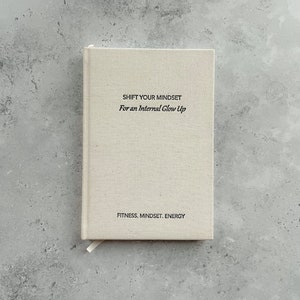 May include: A beige linen journal with the text "SHIFT YOUR MINDSET For an Internal Glow Up" and "FITNESS. MINDSET. ENERGY" printed on the front cover.