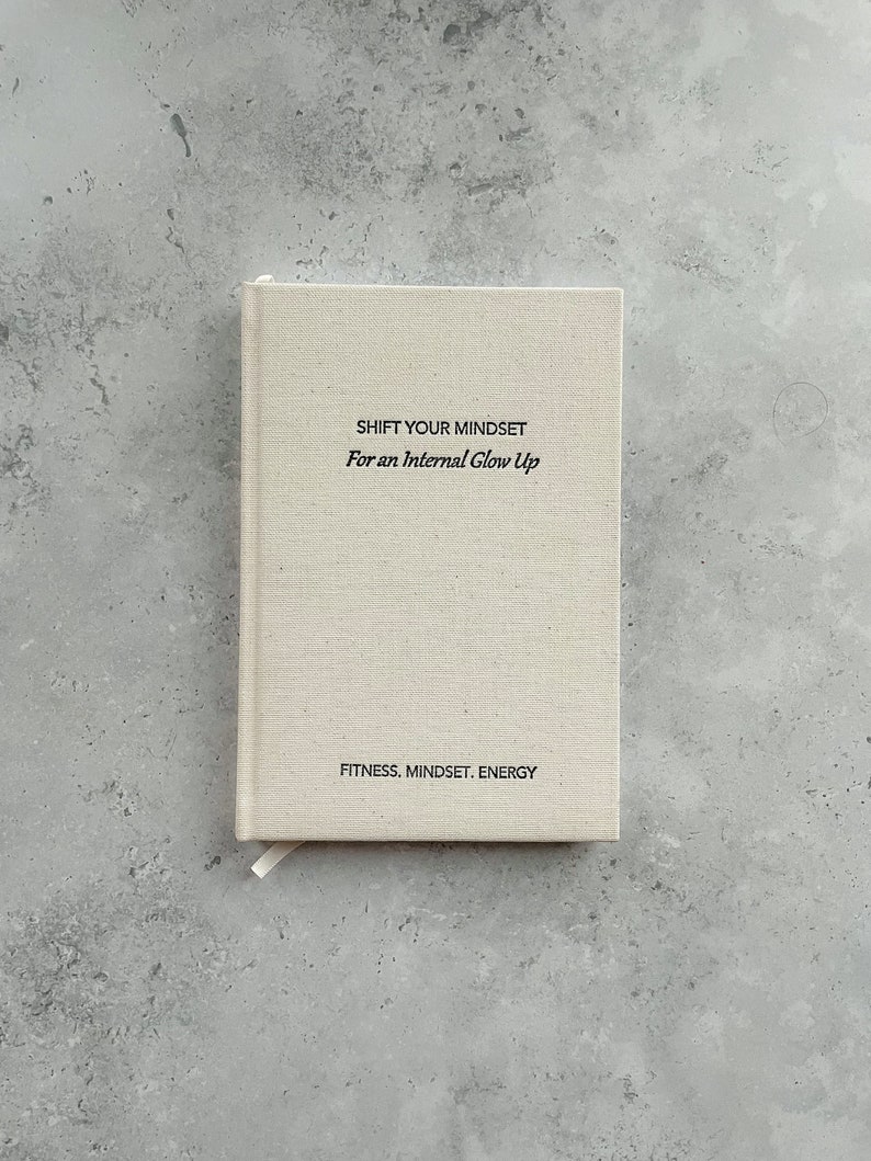 May include: A beige linen journal with the text "SHIFT YOUR MINDSET For an Internal Glow Up" and "FITNESS. MINDSET. ENERGY" printed on the front cover.