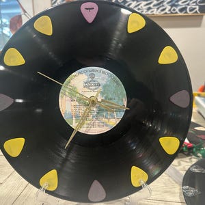 Pode incluir: Um relógio de vinil preto com mostrador e ponteiros dourados. O relógio é decorado com 12 palhetas de guitarra de várias cores, incluindo amarelo, roxo e cinza. O rótulo do disco diz "Park Home of Walter Back Record".