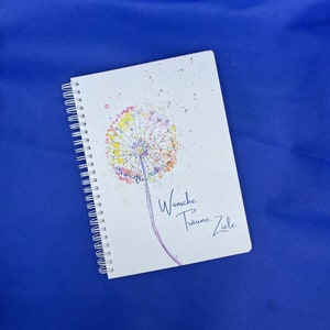 Könnte beinhalten: Ein weißes Spiralblock mit einer Aquarell-Illustration eines Löwenzahns mit bunten Blütenblättern und dem Text "Wünsche, Träume, Ziele".