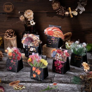 May include: Assortment of clear stickers featuring various floral arrangements and botanical designs. The stickers are displayed on black triangular stands with a floral pattern. The text on the stands reads "Floral Mystery", "Botanical Menu", "Tulips", "Nightbloom Minimal", and "Roses".