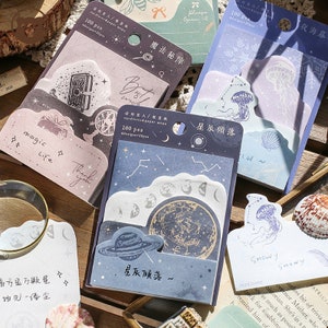 May include: A set of four packs of decorative memo pads with a celestial theme. Each pack features a different design with a blue, purple, or pink background and gold accents. The designs include stars, planets, moons, and constellations. The memo pads are labelled with the text "星辰傾落" which translates to "Stars Falling".