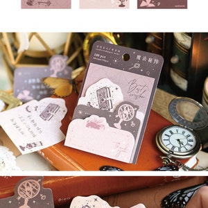 May include: A pack of 100 decorative stickers with a celestial theme. The stickers feature a book, a globe, a key, and a bottle. The stickers are in shades of pink, brown, and white. The text on the stickers reads "Best wishes", "Thank you", "Love life", and "Every little sparkle is worth celebrating!"
