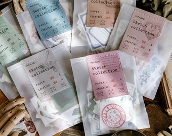 Könnte beinhalten: Fünf durchsichtige Plastiktüten gefüllt mit Vintage-Papieraufklebern. Jede Tüte hat ein Etikett mit der Aufschrift "MO-CARD Sketch collection" mit einem Design eines Vintage-Stempels. Die Etiketten enthalten auch den Text "PAPER 100..." und "CREATIVE 100..." mit einem Häkchen-Symbol.