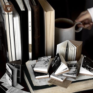 以下が含まれることがあります： 「Black and White」という文字が書かれた、小さく、黒と白のノートの集まり。ノートは積み重ねられており、本とコーヒーカップに囲まれています。