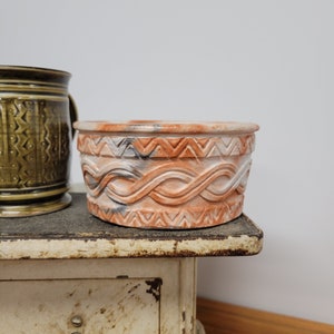 Puede incluir: Una maceta de terracota con un esmalte blanco y naranja. La maceta tiene un patrón decorativo de líneas onduladas y triángulos alrededor del borde superior.