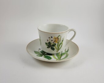 May include: A white porcelain teacup and saucer set with a floral design featuring yellow and green flowers. The cup has a gold rim and the saucer has a gold edge.