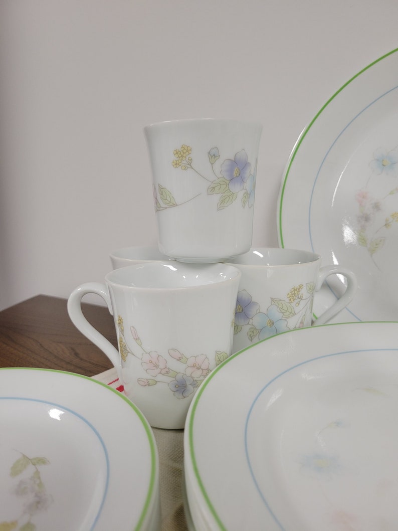 May include: A set of white ceramic mugs and plates with a floral design in shades of blue, pink, and green. The mugs have a handle and the plates have a green and blue rim.