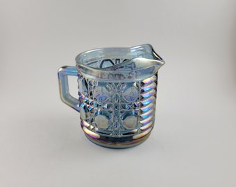 May include: Iridescent blue glass creamer with a handle and spout. The creamer has a textured, geometric pattern with a series of circles and diamond shapes. The glass reflects a rainbow of colours, including purple, green, and gold.