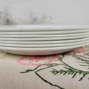 May include: A stack of white ceramic plates with a slight green tint around the edges. The plates are stacked on a white fabric with a floral embroidery design.
