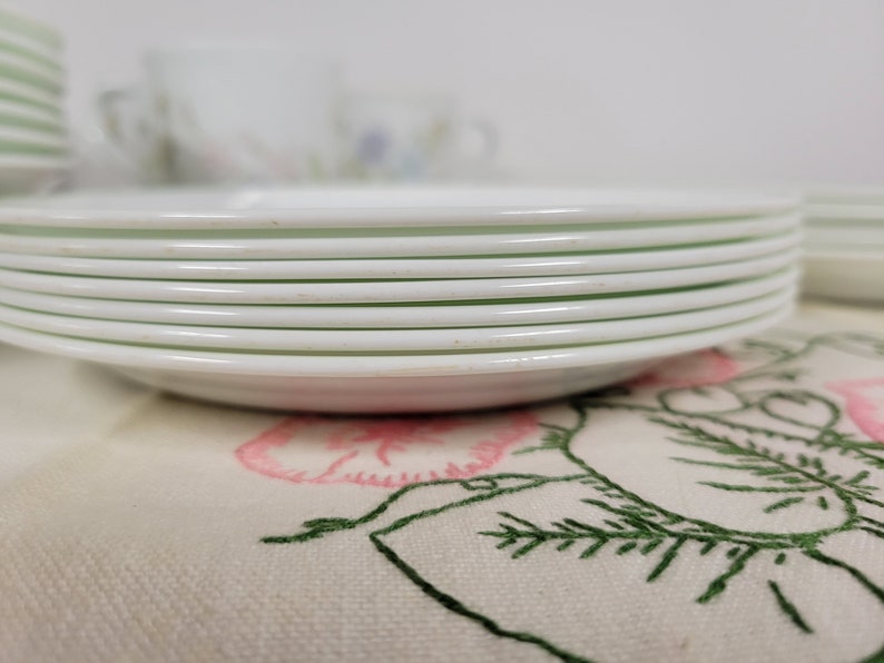 May include: A stack of white ceramic plates with a slight green tint around the edges. The plates are stacked on a white fabric with a floral embroidery design.