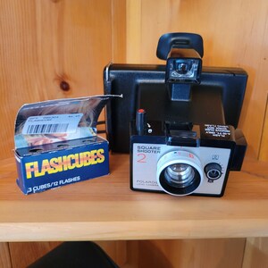 May include: A black Polaroid Square Shooter 2 camera with a flashcube and a pack of flashcubes. The camera has a white lens and a black and orange label that says "Square Shooter 2" and "Polaroid Land Camera".