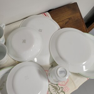 May include: A set of white Corelle dinnerware with a floral pattern. The set includes a large dinner plate, a smaller salad plate, a teacup, and a saucer. The plates have a green rim and the teacup has a green and pink floral design.