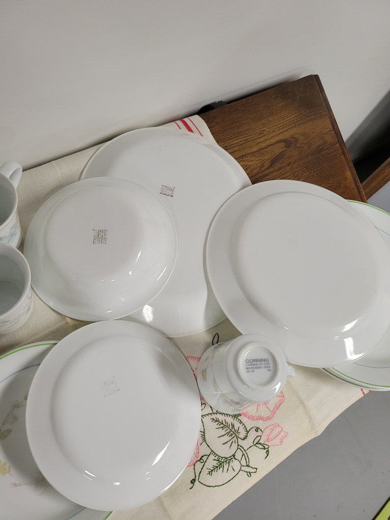 May include: A set of white Corelle dinnerware with a floral pattern. The set includes a large dinner plate, a smaller salad plate, a teacup, and a saucer. The plates have a green rim and the teacup has a green and pink floral design.