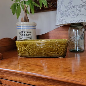 Puede incluir: Una maceta de cerámica verde con un patrón de veta de madera. La maceta está sentada sobre una superficie de madera. El texto "Compliments of Leonard W. McClain 1333 S. 17th St. PHILA. DISTRIBUTOR OF McClain's" está impreso en la maceta.