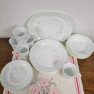 May include: A set of white Corelle dinnerware with a floral pattern and green trim. The set includes a large oval platter, four mugs, four dinner plates, and a set of bowls.