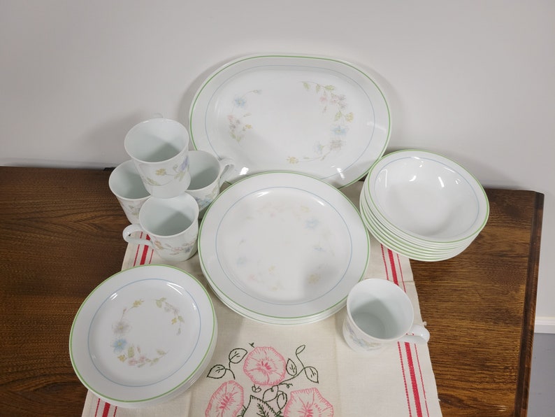 May include: A set of white Corelle dinnerware with a floral pattern and green trim. The set includes a large oval platter, four mugs, four dinner plates, and a set of bowls.
