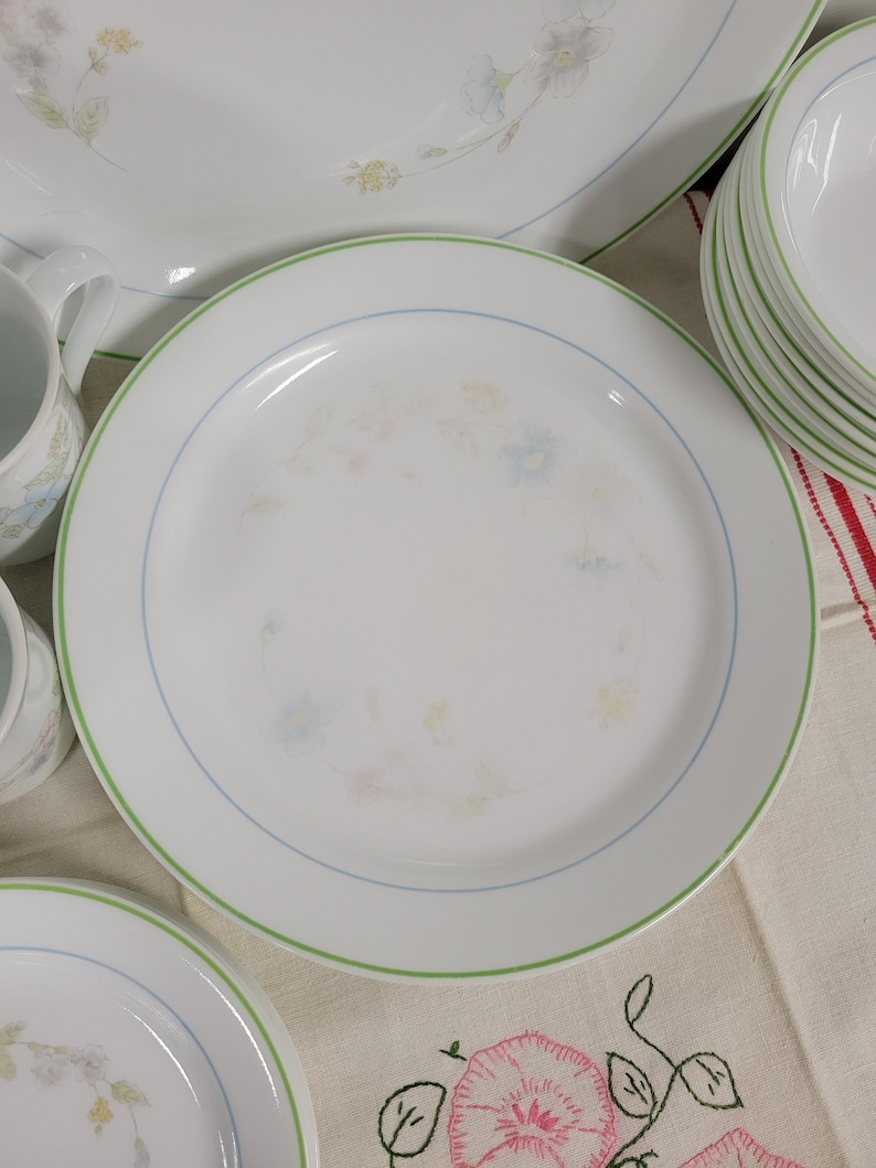 May include: A set of white ceramic dinnerware with a green and blue floral pattern. The plates have a green rim and a blue floral design in the center.
