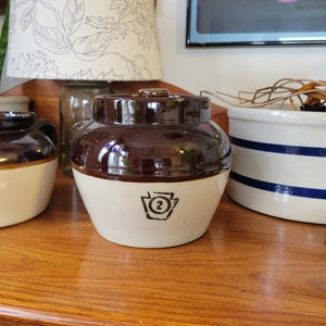 Puede incluir: Un tarro de cerámica marrón y blanco con tapa. El tarro tiene una parte superior marrón oscuro y una base blanca con el número 2 negro dentro de una forma de clave. Otros tarros son visibles en el fondo.