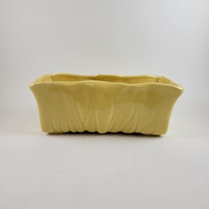 May include: A yellow ceramic planter with a rectangular shape and a textured, wavy design. The planter is made of glazed ceramic and has a smooth, glossy finish.