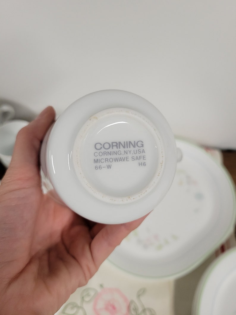 May include: White ceramic mug with the text "CORNING CORNING, NY, USA MICROWAVE SAFE 66-W H6" on the bottom.