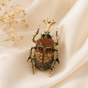 Puede incluir: Un broche de escarabajo en tono dorado con un cuerno prominente y seis patas. El cuerpo del insecto está incrustado con un degradado de pedrería que va desde el ámbar y naranja en la parte superior hasta el marrón oscuro y gris en la base.