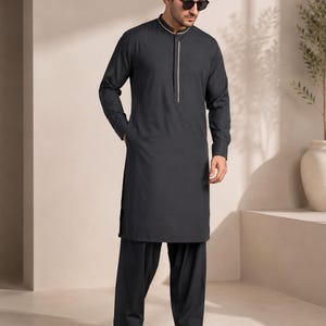 Könnte beinhalten: Ein dunkelanthrazitfarbener, langärmeliger Kurta mit Stehkragen und vertikalem, weißem Zierstreifen entlang der Knopfleiste, kombiniert mit einer passenden, locker sitzenden Hose und schwarzen, offenen Sandalen, getragen mit einer dunklen Sonnenbrille.