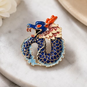 Puede incluir: Un broche de metal dorado con forma de dragón enroscado con escamas de esmalte azul, una cresta naranja y detalles de crin color crema. El dragón tiene la boca abierta con dientes visibles y un cuerpo curvo que forma un diseño circular.