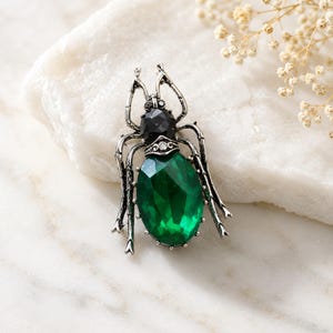 Puede incluir: Un broche de insecto en tono plateado con una gran piedra preciosa ovalada de color verde esmeralda para el cuerpo y una piedra negra redonda más pequeña para la cabeza. El marco de metal tiene patas texturizadas y un pequeño cristal transparente.